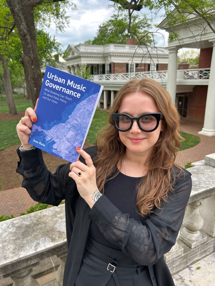 A person dressed in black, with red hair and wearing glasses holds a book with a blue cover that says: Urban Music Governance: What Busking Can Teach US about Data, Policy and Our Cities, Jess Reia. The University of Virginia historical lawn is in the background. It is daytime.