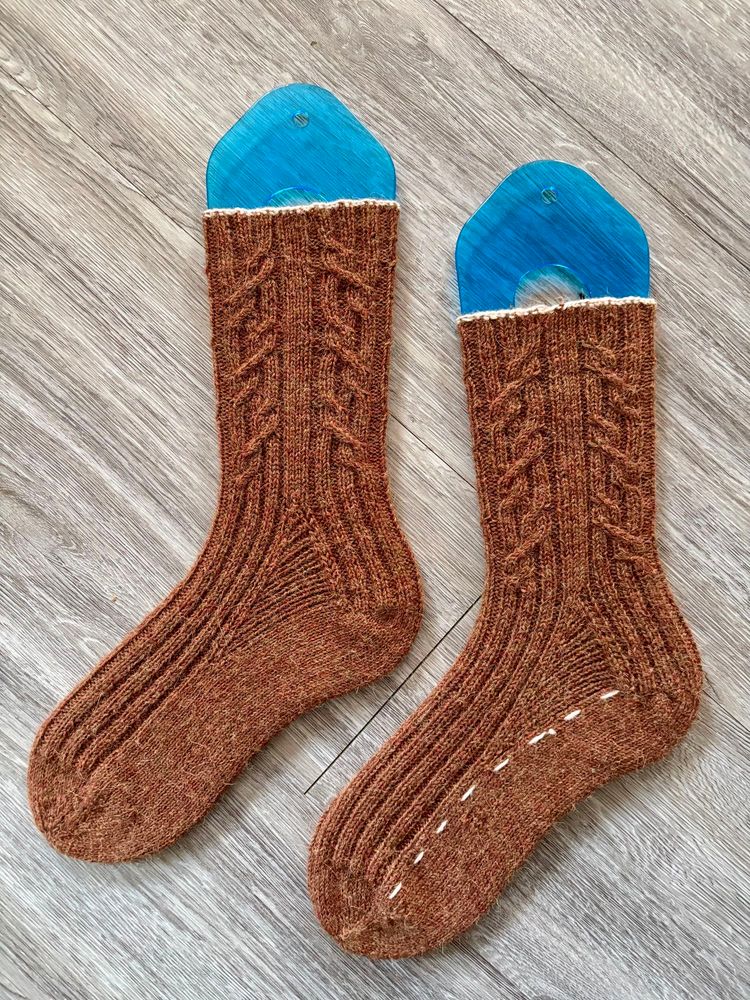 Brown hand knit socks with a cable pattern on the leg and a white thin cuff on blue sock blockers and a grey background.
