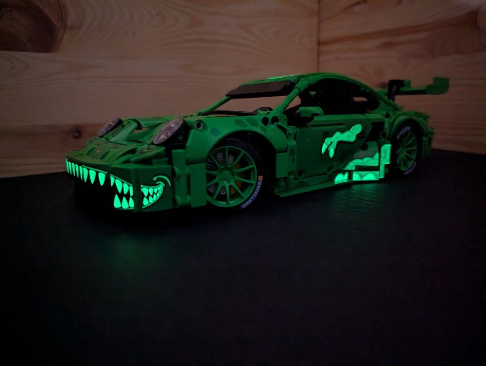 A brick built (not Lego) Porsche 911 GT3-R with the "Rexy" livery, kind of depicting a T-Rex. Shown from the front left in the dark, to show off the glow in the dark stickers. Teeth, claws and part of the body are glowing!