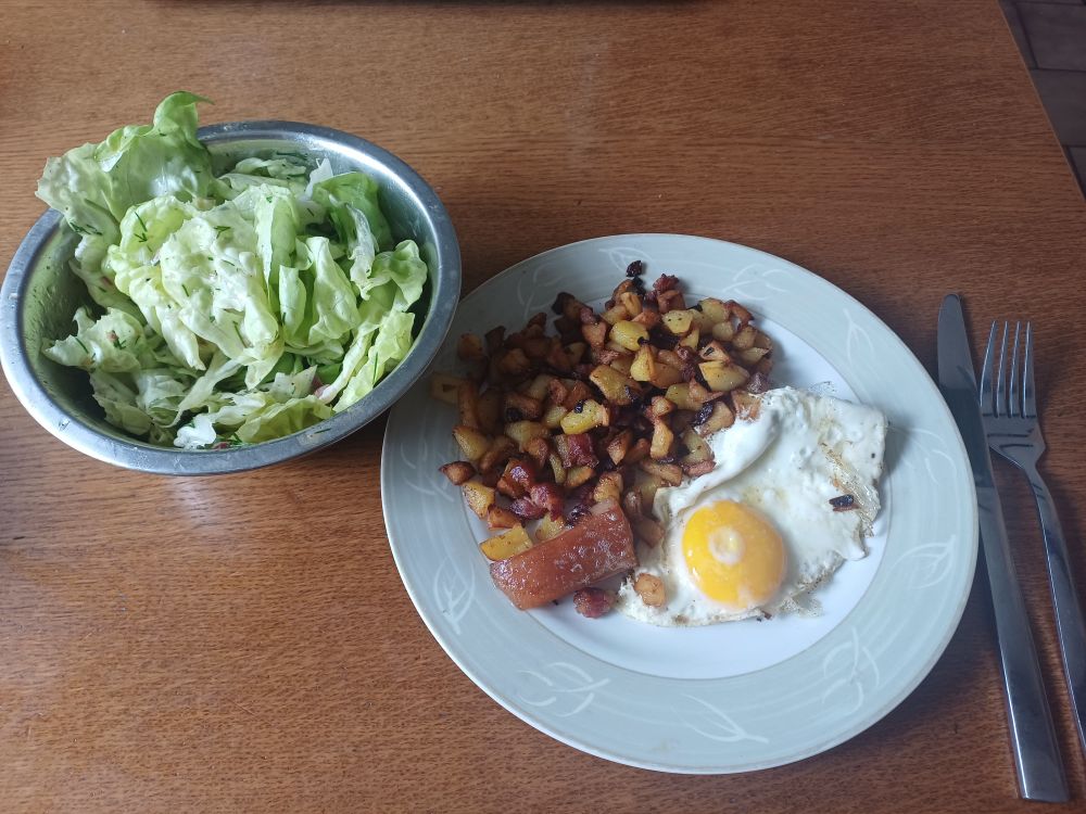 Teller mit Bratkartoffeln und Spiegelei dazu Schüssel mit grünem Salat.
