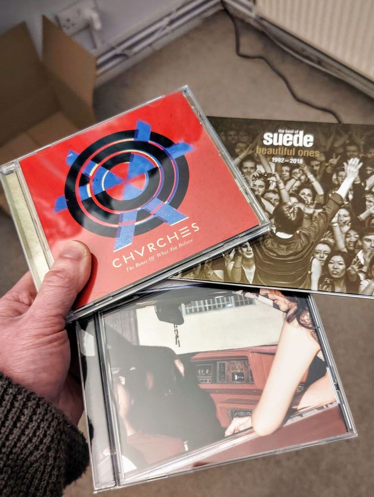 Hand holding three CD album cases: 'The Bones Of What You Believe' by CHVRCHES, 'Beautiful Ones - The Best of Suede 1992-2018' by Suede and 'Lonely People With Power' by Deafheaven.