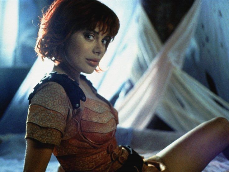 Xenia Seeberg as Xev Bellringer in the Lexx TV series. A woman with short bobbed red hair wearing a short-sleeved tunic seemingly made of reptile scales with leather shoulder straps. She reclines in front of a backdrop of flowing white veils.