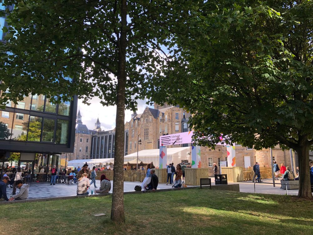A set of modern and Victorian office buildings beside a square. Tents and banners and lots of people show it’s the Edinburgh Book Festival