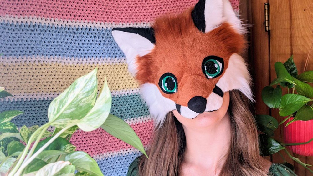 a red fox therian mask with green eyes being worn by a critter with long brown hair. she is standing in front of a horizontal striped crochet blanket wall hanging and there are potted pothos plants hanging on either side of her.