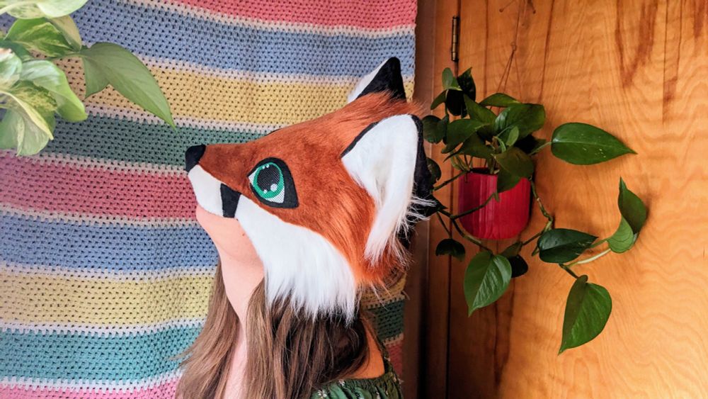 a red fox therian mask with green eyes being worn by a critter with long brown hair. she is standing in front of a horizontal striped crochet blanket wall hanging, sniffing one of the potted pothos plants that are hanging on either side of her.