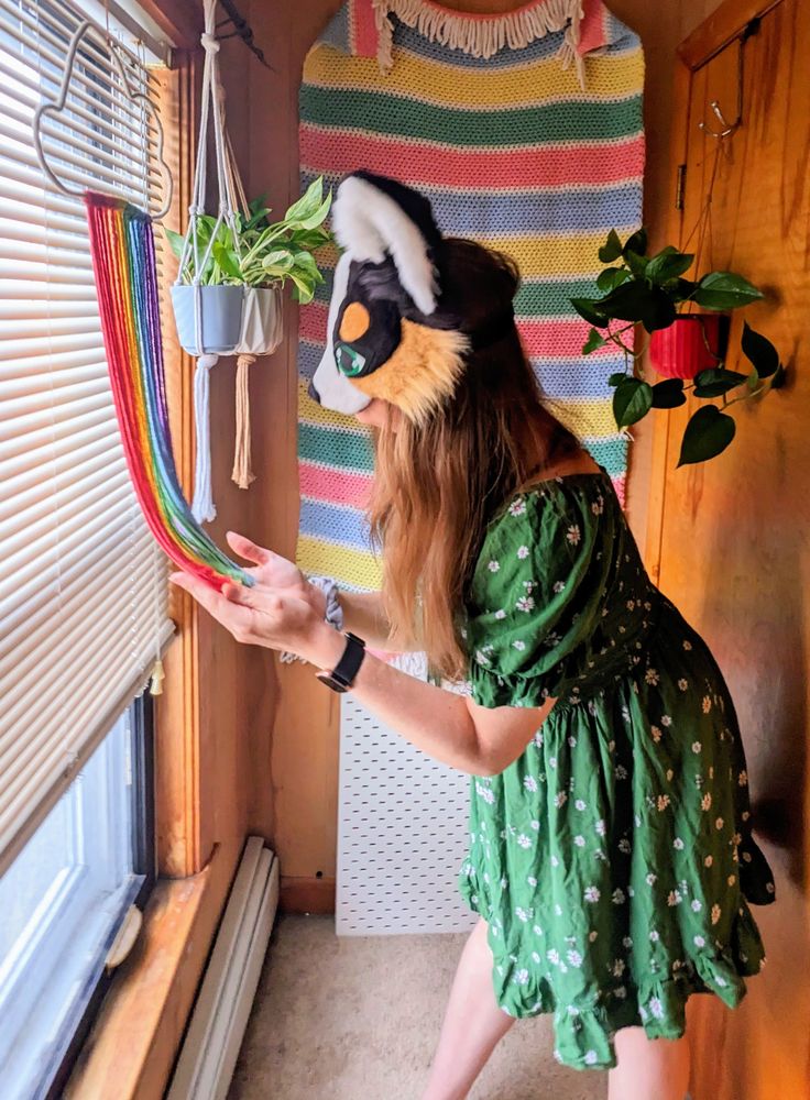 a black tri color austrialian shepherd therian mask with green eyes being worn by a critter with long brown hair and a green dress. she is standing in front of a horizontal striped crochet blanket wall hanging and there are potted pothos plants hanging on either side of her, she is holding the rainbow tassels of a pride wall hanging in front of a window with the blinds mostly down.