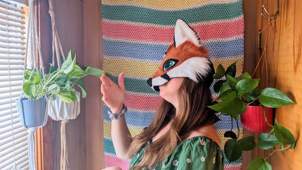 a red fox therian mask with green eyes being worn by a critter with long brown hair. she is standing in front of a horizontal striped crochet blanket wall hanging, holding a paw up to one of the potted pothos plants that are hanging on either side of her.