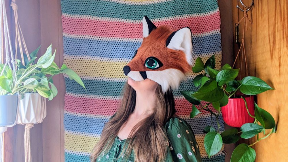 a red fox therian mask with green eyes being worn by a critter with long brown hair. she is standing in front of a horizontal striped crochet blanket wall hanging and there are potted pothos plants hanging on either side of her.