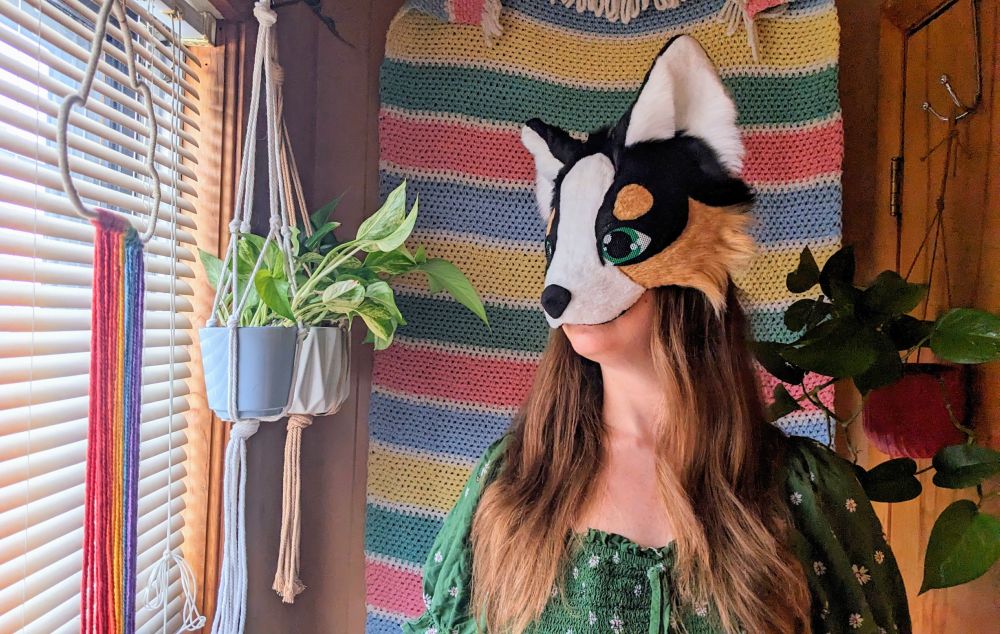 a black tri color austrialian shepherd therian mask with green eyes being worn by a critter with long brown hair. she is standing in front of a horizontal striped crochet blanket wall hanging and there are potted pothos plants hanging on either side of her.
