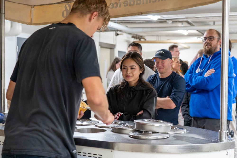 A young woman in a black jacket stands at an ice cream cart, watching as the server scoops ice cream. Behind her, a group of people including a man in a navy hoodie and another in glasses patiently wait their turn.