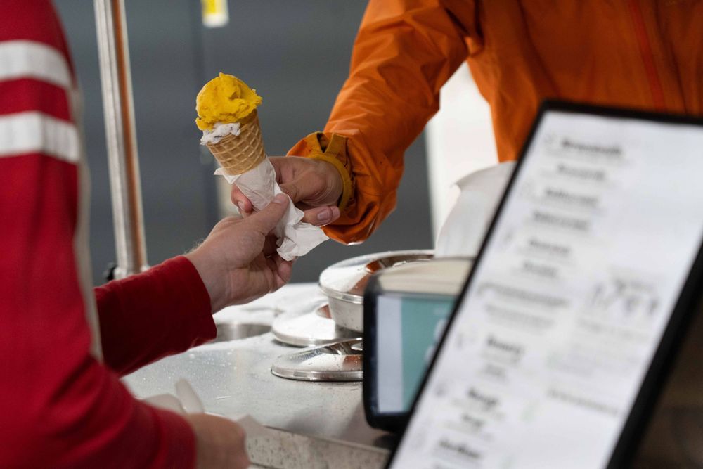 A close-up of an ice cream cone being handed to a person. The cone has two scoops: one bright yellow and one white with dark specks. The server in an orange jacket passes it across the ice cream cart counter, which has a menu and ice cream containers visible.
