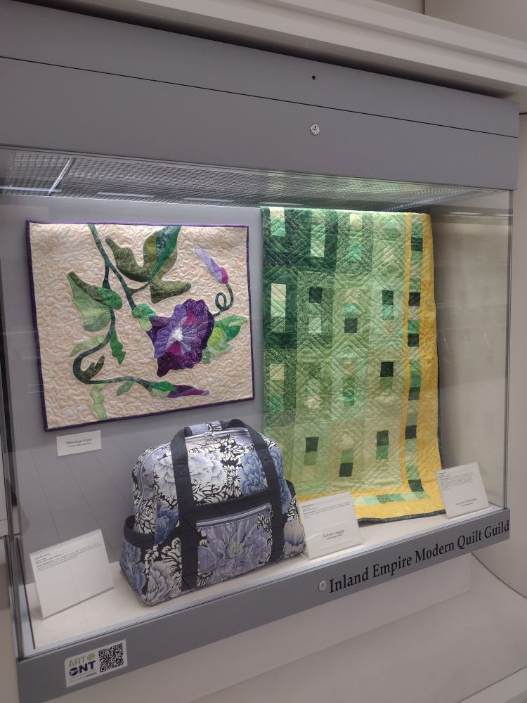 Display case at Ontario Airport displaying a large green and gold ombre quilt, a quilted travel bag, and a quilted wall hanging of a flower.