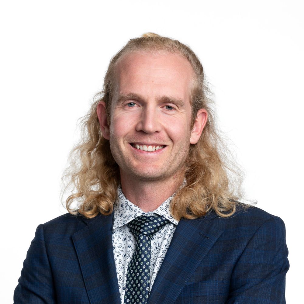 Image of Alex Baker in a navy blue plaid suit with a white button down shirt that has a small detailed pattern on it with dark blue. He is wearing a satin tie that is dark blue with a gold pattern on it. He is wearing a relaxed smile and has shoulder length curly blond hair that is half tied back.