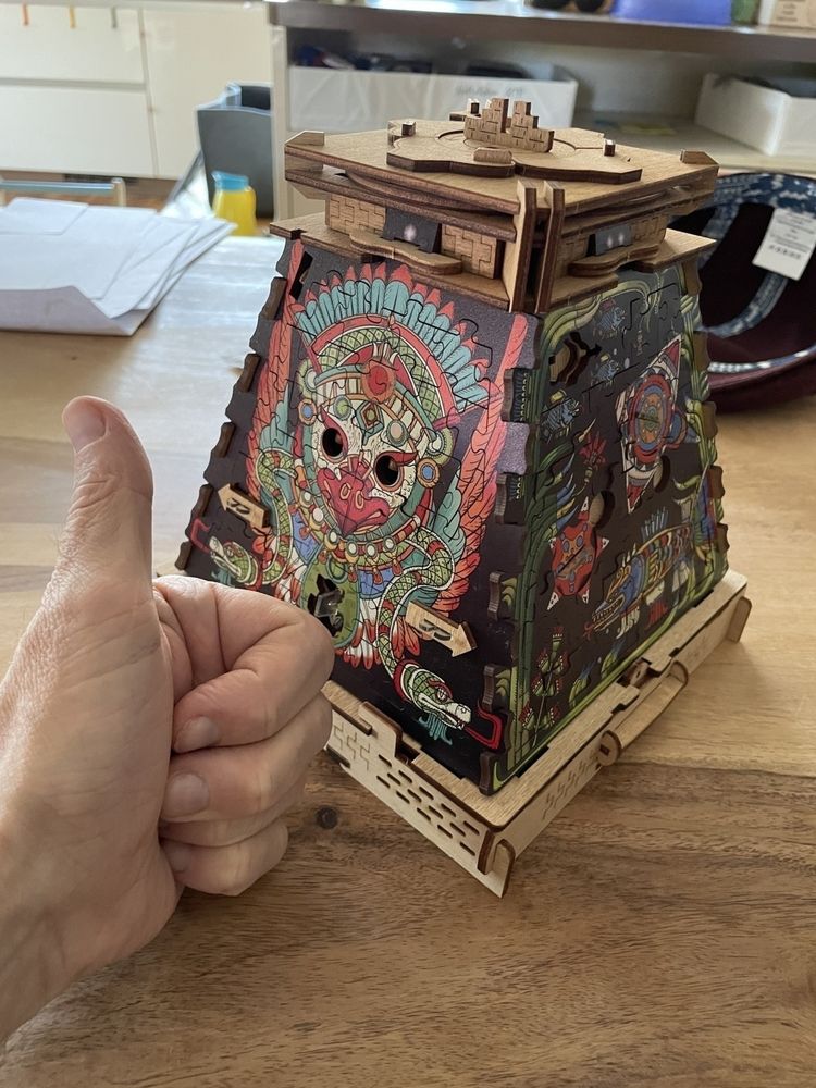 Auto-generated description: Ein Daumen hoch wird neben einem detailreichen Holzpuzzle in Form einer dekorativ gestalteten Pyramide gezeigt.