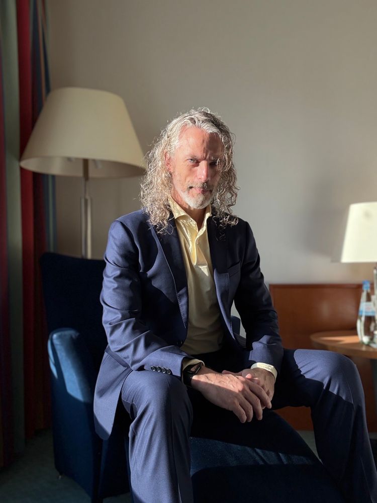 Photo of white man with long curly hair in navy suit & light yellow polo shirt 