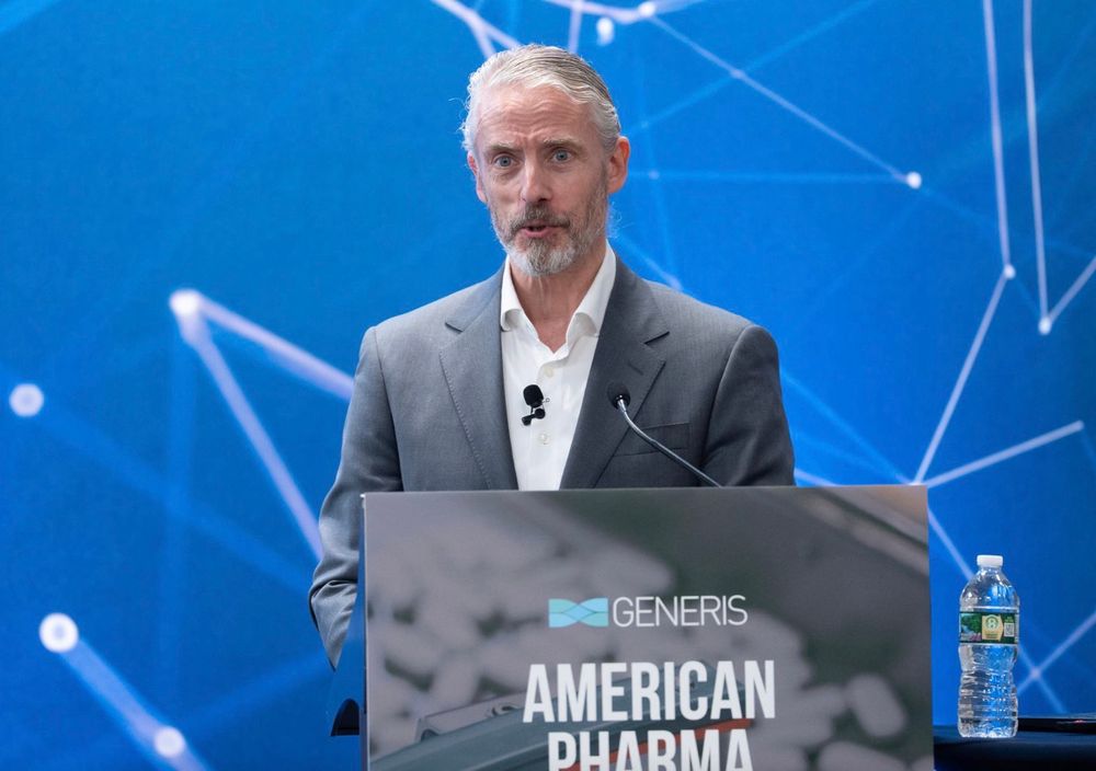 Photo of white man in gray suit & white dress shirt standing a lectern 