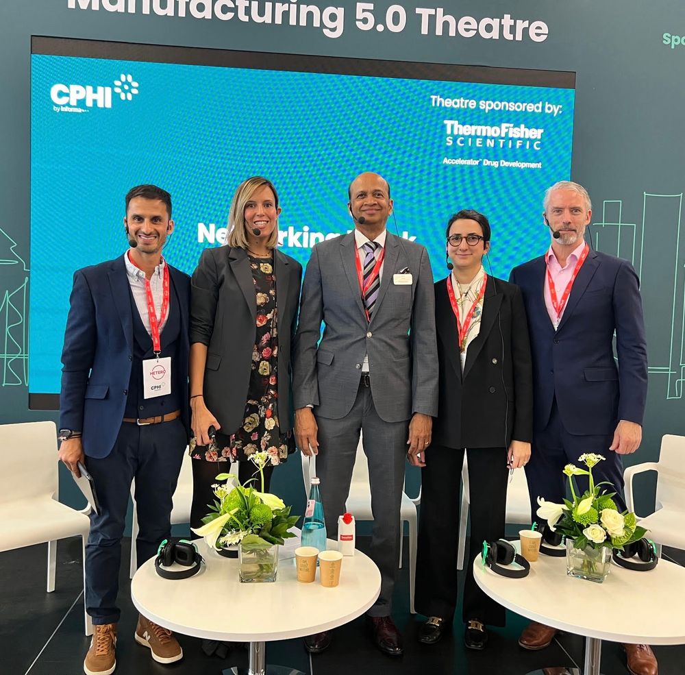 Photo of 5 people who standing & smiling at camera after a panel they participated in. They all have ear-wraparound mics