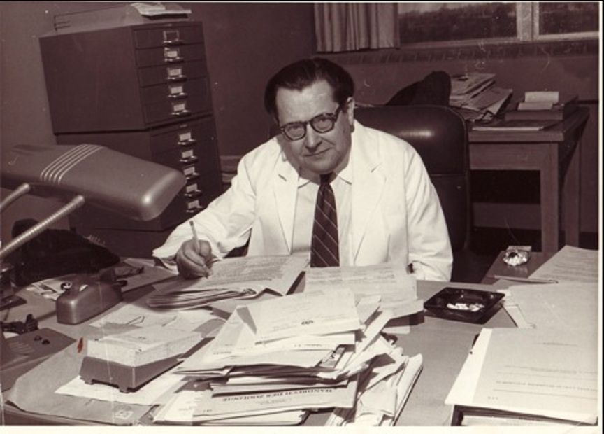 Ludwig von Bertalanffy sitting at his desk - looking very 1950 ish...  with a pencil in hand, white lab coat and tie, slicked back hair, messy stack of papers from a typewriter, ashtray on desk, old style phone, stacks of file cabinets in the back...  Austrian biologist and systems theorist (1901–1972), 1958, Mt Sinai Hospital, LA, USA. Photo courtesy of BCSSS, Bertalanffy Center for the Study of Systems Science, Vienna  - from https://link.springer.com/chapter/10.1007/978-3-658-42117-5_4
