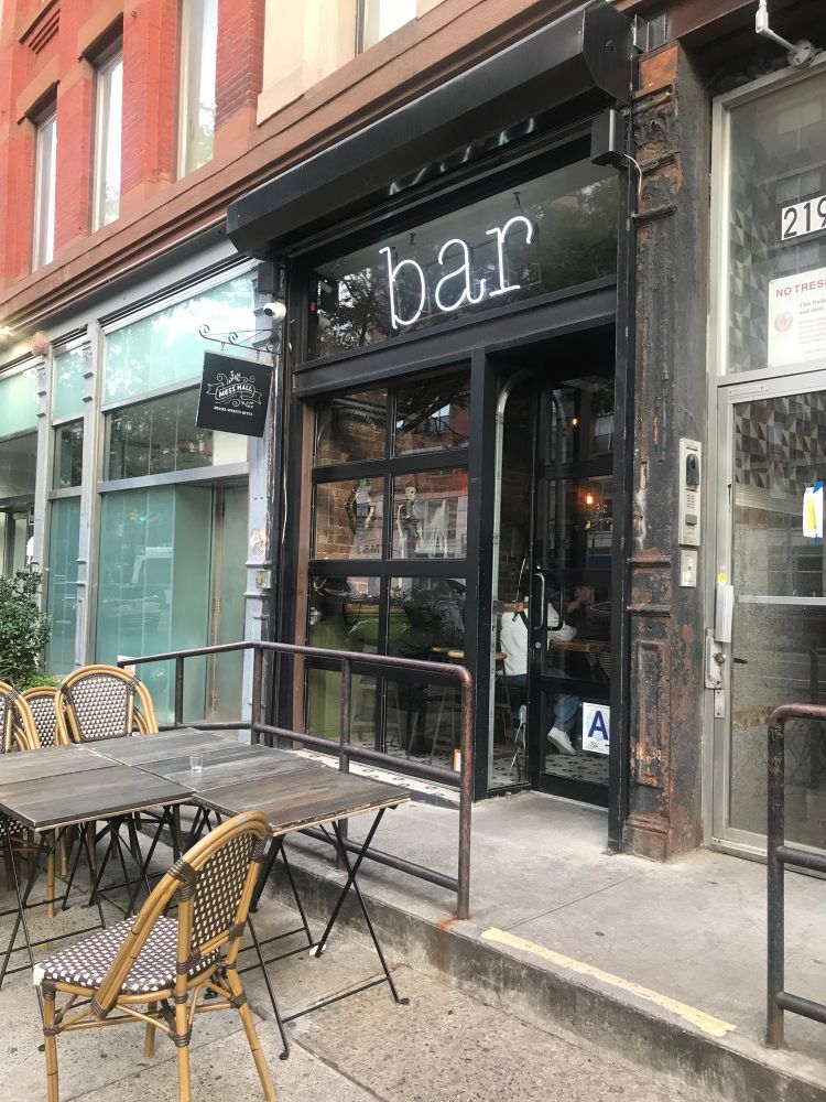 The outside of a bar with a black sign that says BAR in white neon lights and some tables and chairs outside 
