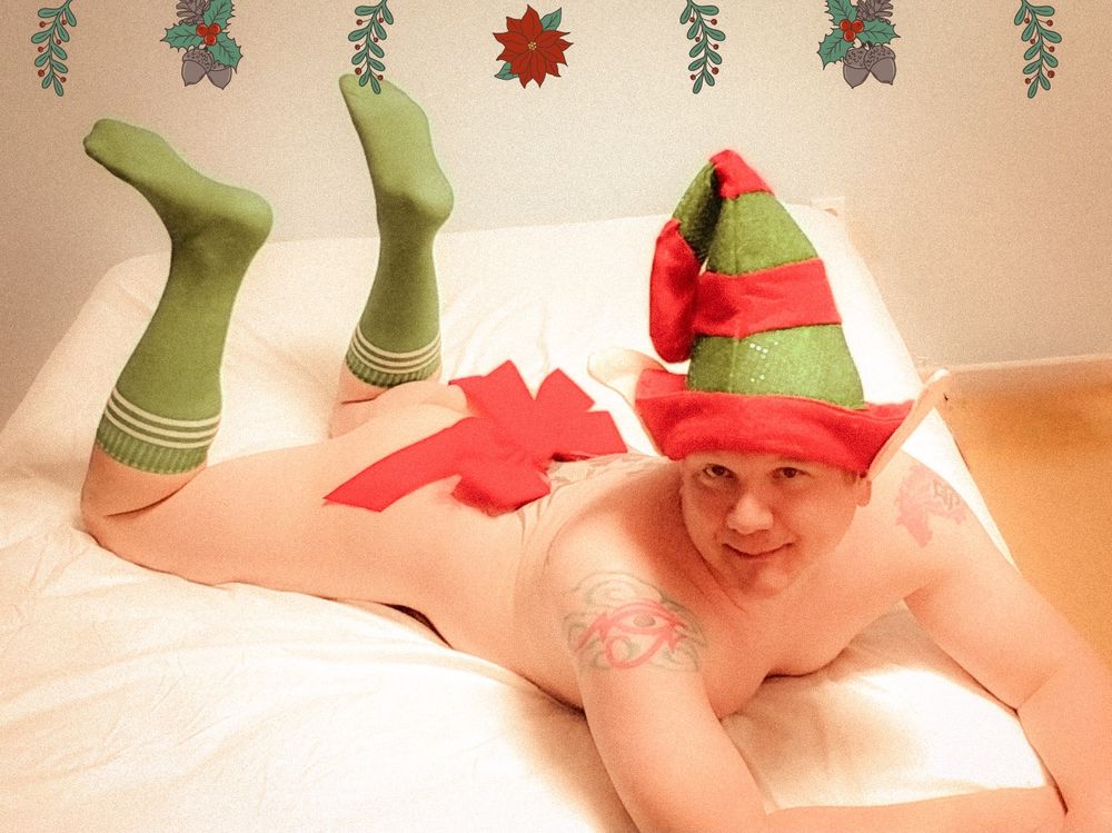 Image is of me mostly naked except for an elf hat and green socks laying on the bed. The photo theme is holiday elf. I'm looking at the camera. I have a red bow right above my butt as I'm laying down.
