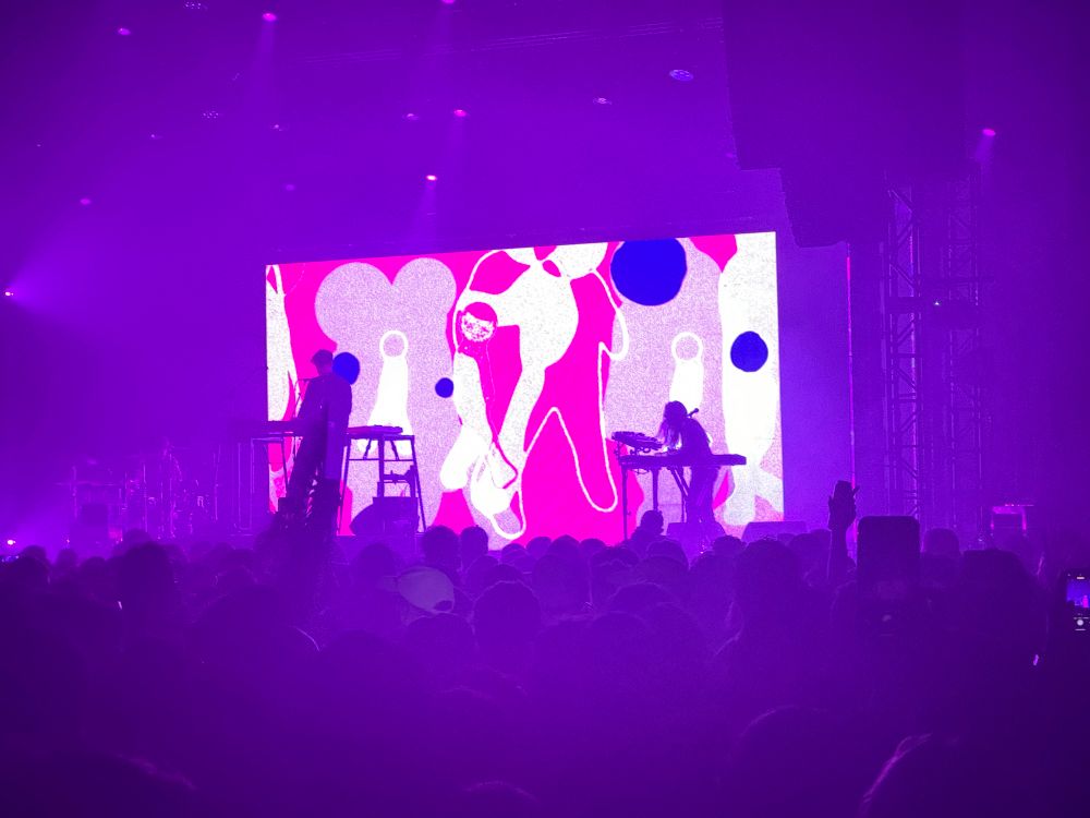 Barry Can’t Swim on stage with purple lights and a video screen showing animations of pink and white figures.