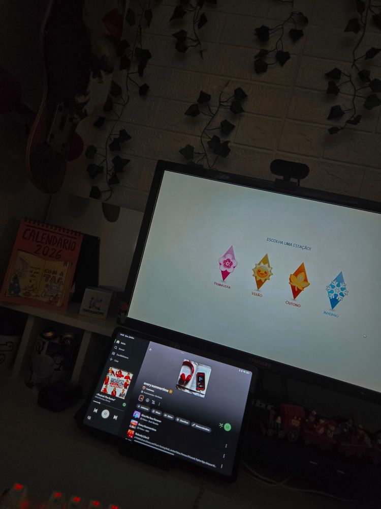 Tela de computador exibindo opções de estações do ano no The Sims, com um tablet abaixo tocando música no Spotify, ao redor decoração com plantas artificiais e alguns enfeites.