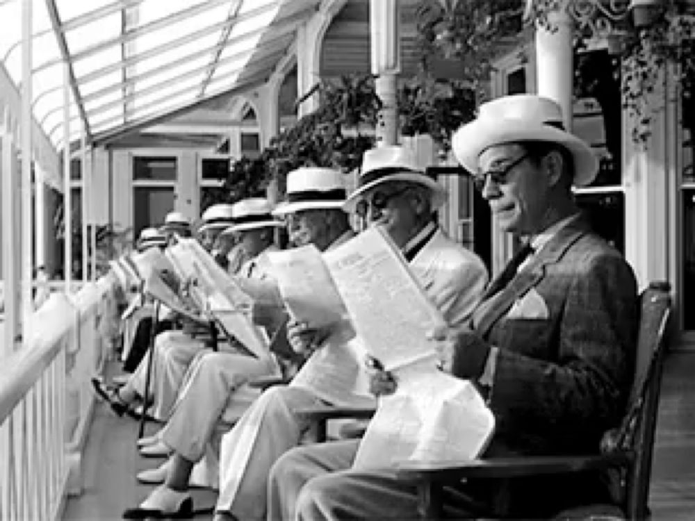 a black and white photo of a group of men sitting on a porch reading newspapers