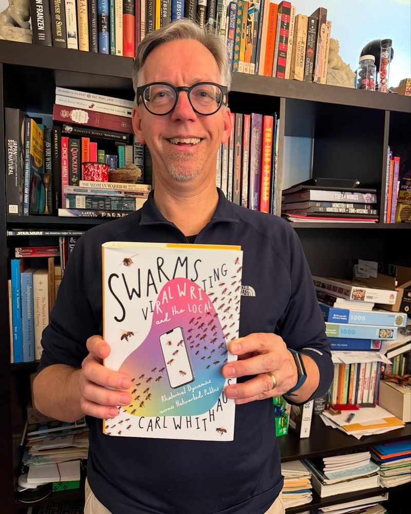 Carl Whithaus, a middle-age man, holds a hardback book titled Swarms, Viral Writing, and the Local. He stands in front of a messy bookshelf.