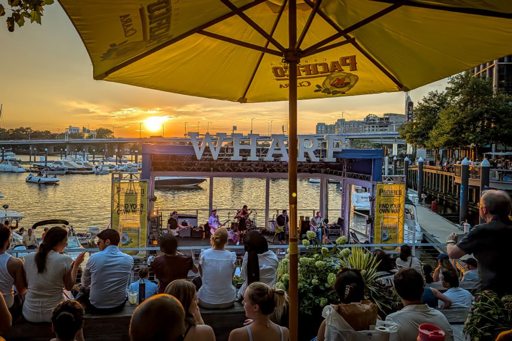 A wide-angle, eye-level shot captures a group of people enjoying a live music performance at an outdoor venue next to Washington Channel waterway. Yellow-orange sunset in the background. The scene is taking place at the Wharf development in Washington DC including a large "WHARF" sign on the stage.