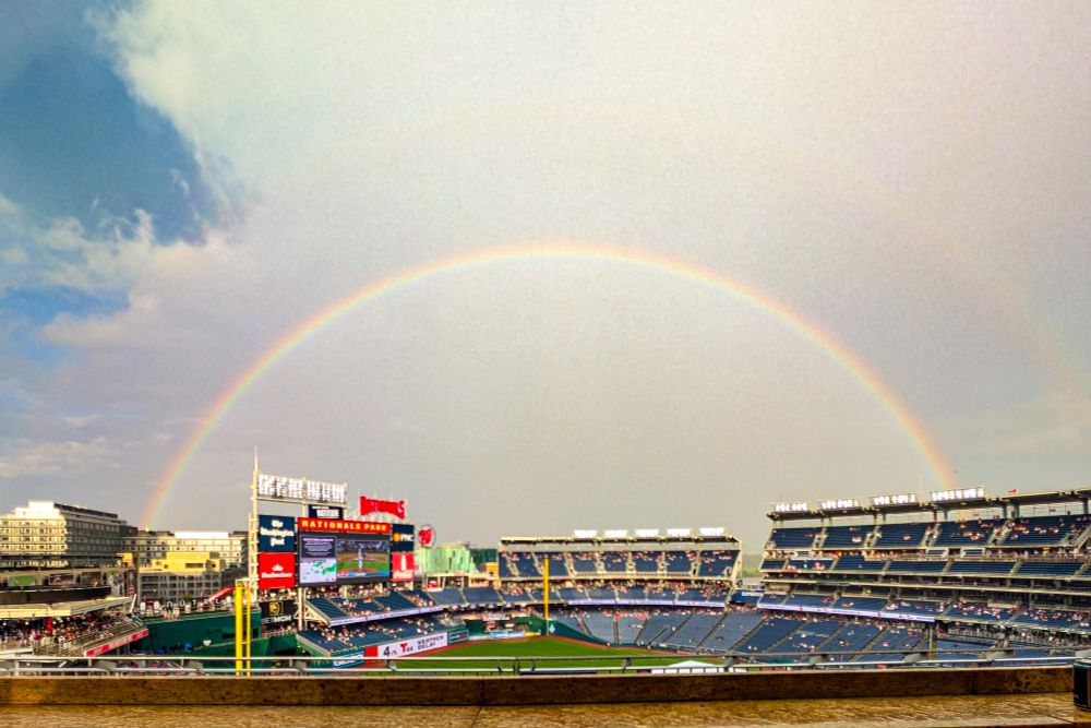 A wide-angle, slightly elevated outdoor shot shows a full, vibrant double rainbow arcing over Nationals Park, home of the Washington Nationals baseball team. The stadium is illuminated under the arch of the rainbow, with the stands sparsely filled with spectators. The sky is partially cloudy, creating a dramatic backdrop. 