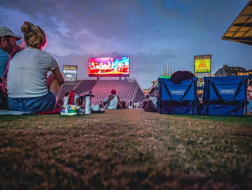 Photo from the ground level of a sports stadium. Empty seats can be seen in the distance next to a large video screen showing a movie. The backs of other people dot the grassy field.