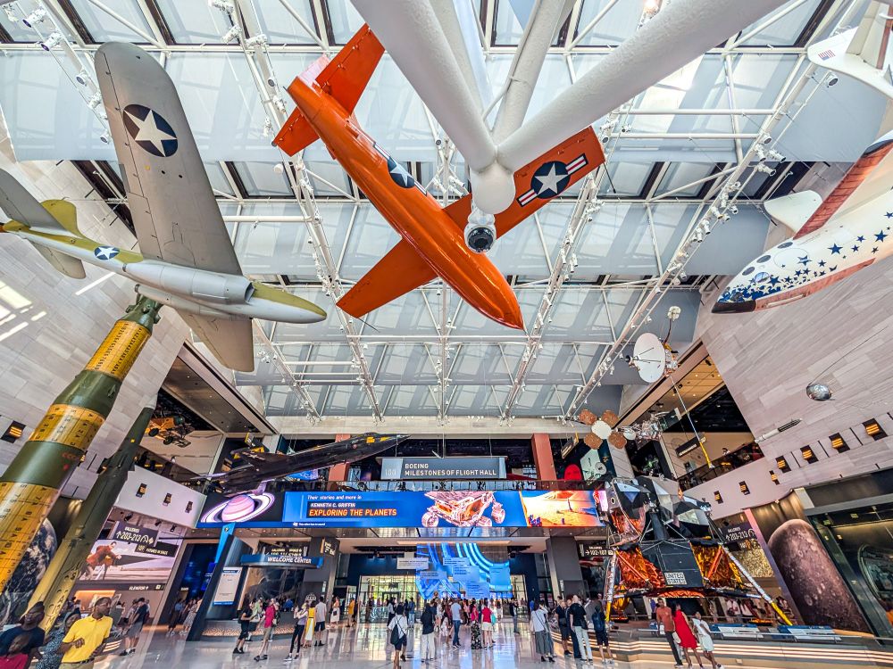 A large, high-ceilinged atrium of the Air and Space museum in Washington DC. The atrium is filled with a variety of suspended aircraft and space vehicles. The ceiling is a grid of glass panels, allowing natural light to flood the space.

The floor is a wide, open space filled with a crowd of people walking around and looking at the exhibits. The walls of the atrium are lined with more displays and information panels. 