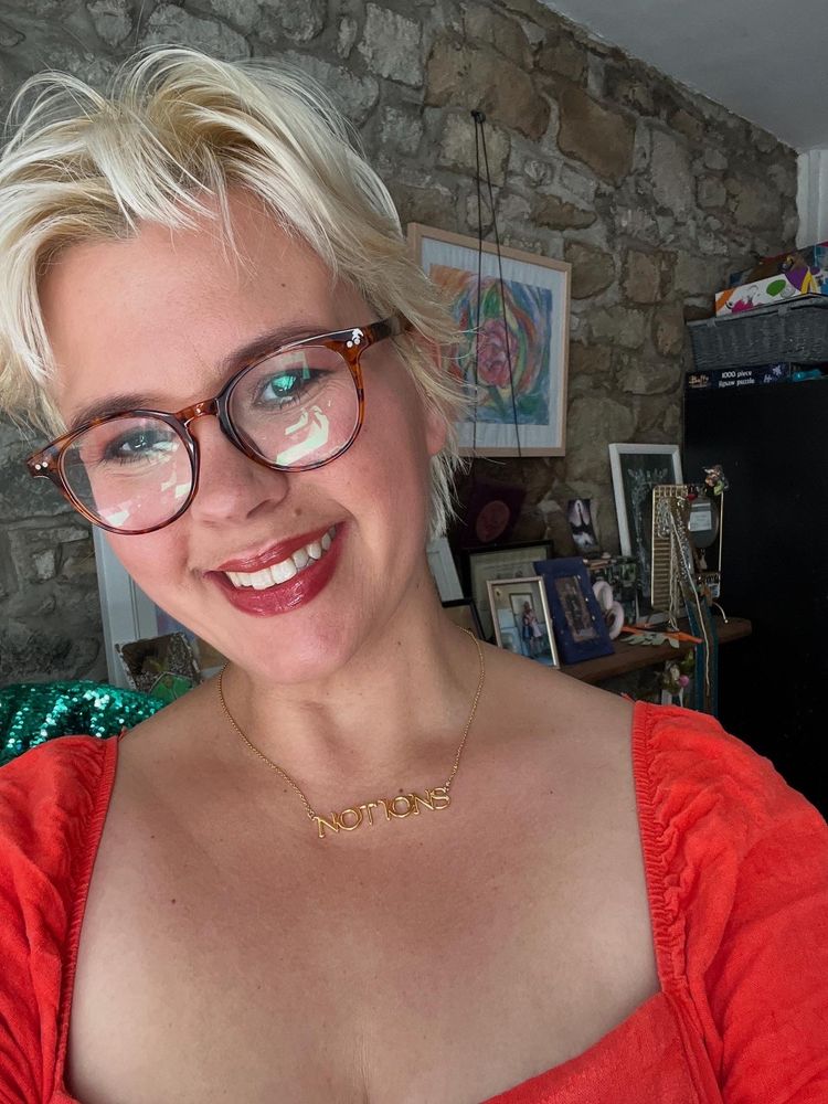 A white woman with bleached hair and brown glasses, wearing an orange dress and a gold necklace that says “notions”