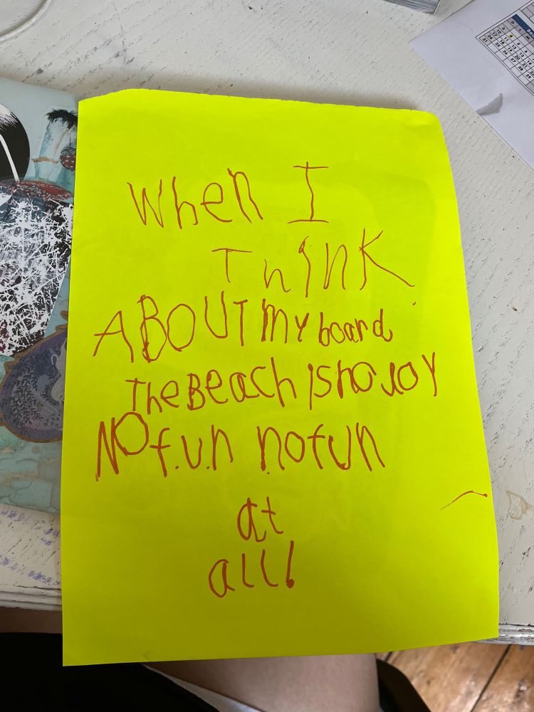 A neon yellow piece of paper with writing in red marker: “when I think about my board, the beach is no joy no f.u.n no fun at all”
