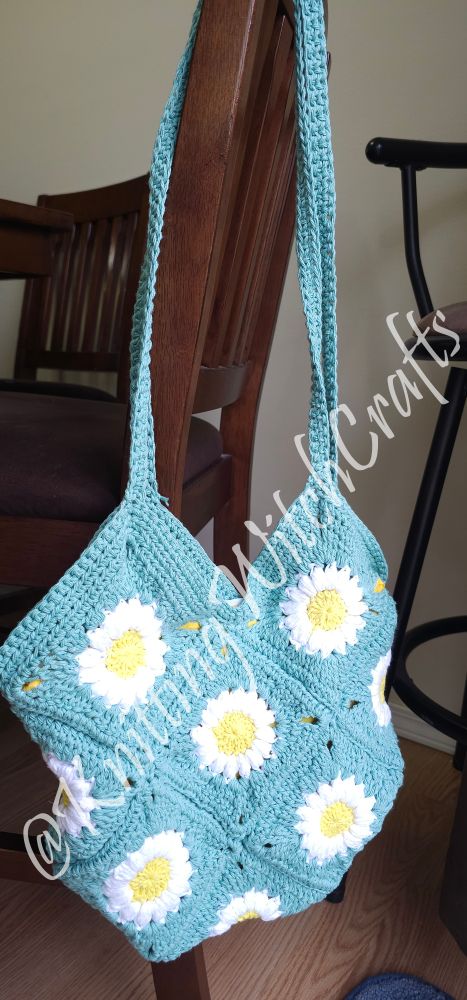 A light teal granny square tote bag hanging over a chair. In the middle of each granny square is a daisy.