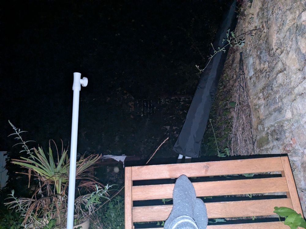 dunkler Himmel, im Schwarz nicht erkennbar:hintere Steinmauer und Baum dahinter/darüber
Rechts im Blitzlicht Steinwand und meine besockten Füße vor Endlehne meiner Gartenbanecke