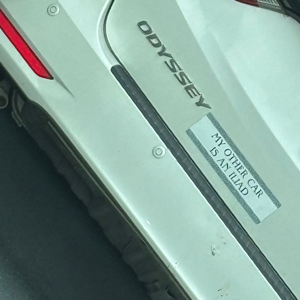 A closeup of a white Honda Odyssey’s bumper. Next to the Odyssey badge is a white bumper sticker with a blue Grecian key border design banding the top and bottom. The text reads MY OTHER CAR IS AN ILIAD. 