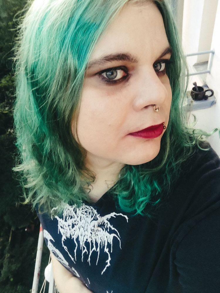 Person with green hair, dark eye makeup, and facial piercings takes a selfie on a balcony, wearing a black Darkthrone shirt.
