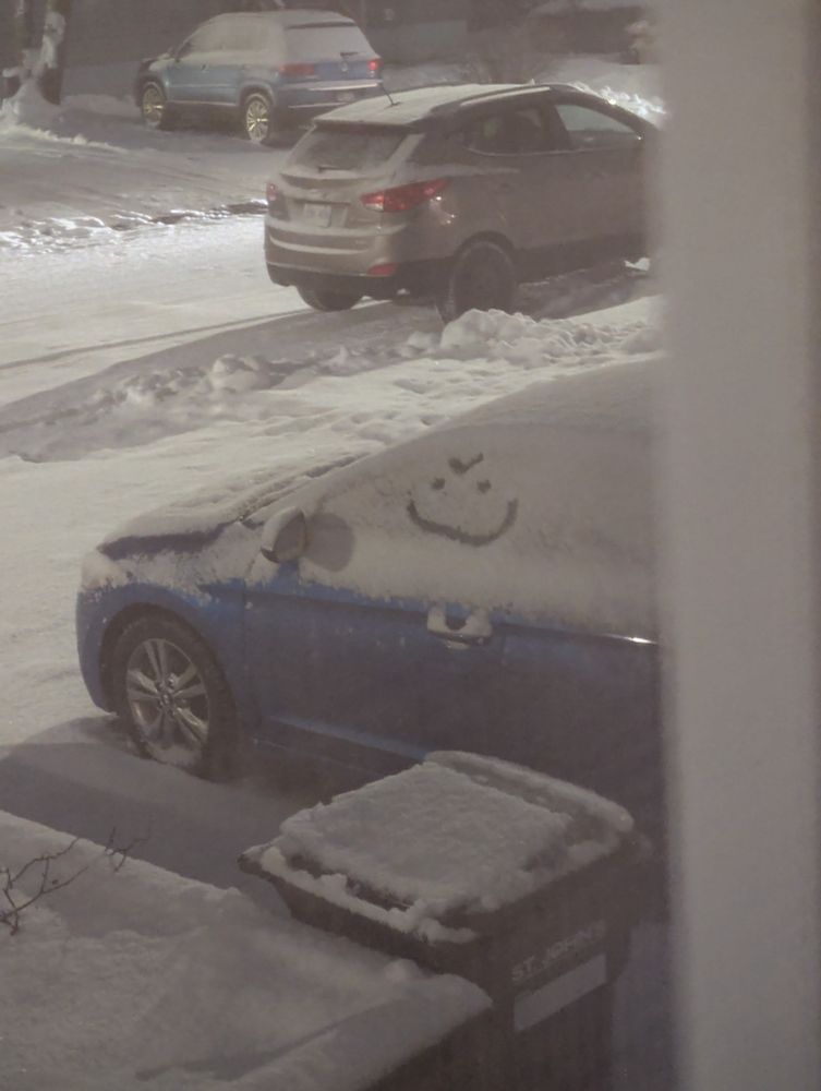 A car covered in snow with an evil smiley face drawn on the driver's window