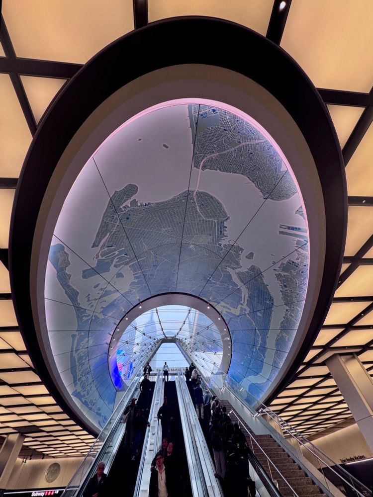 Looking up an exit escalator filled with people. There is a map of nyc on the ceiling above the escalator and lighting panels on the ceiling outside of that. 