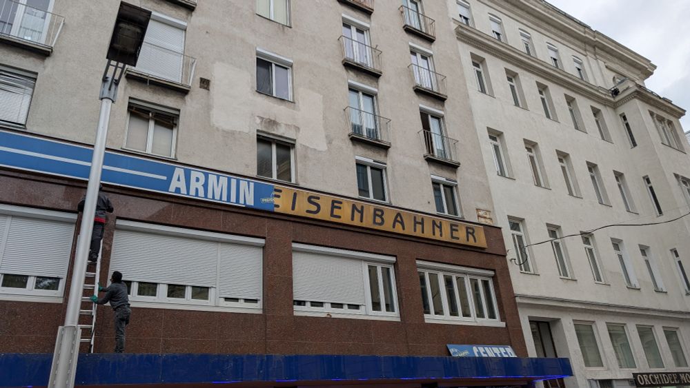 Foto einer Fassade wo ein Schild abmontiert wird. Darunter versteckt sich ein altes Schild "Modehaus Eisenbahner". 