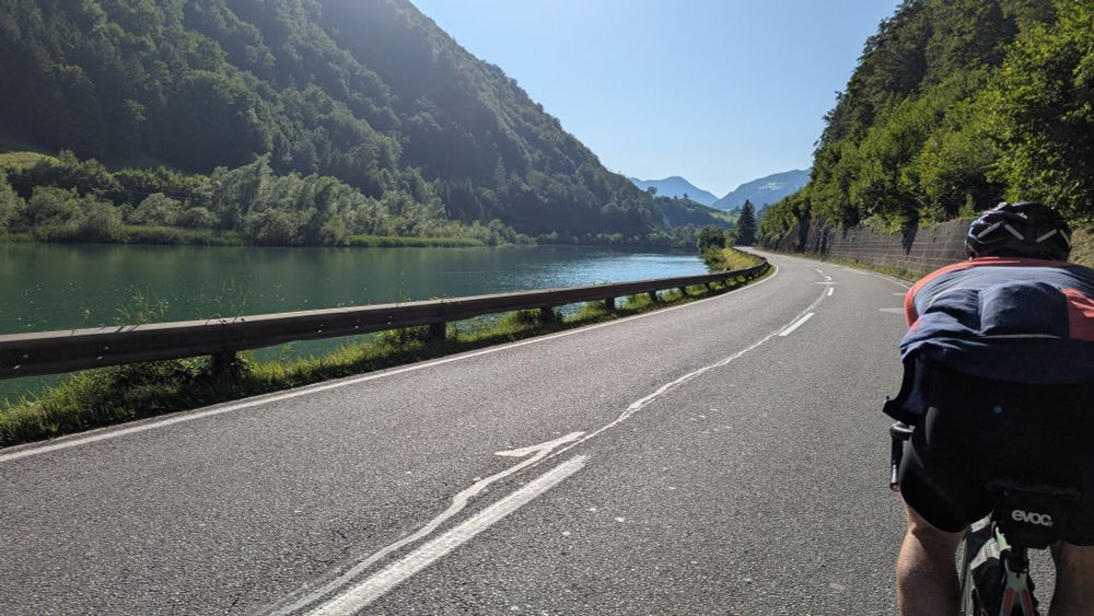 Im Windschatten eines anderen Radfahrers. Die Straße geht entlang eines grün-blauen Flußes. Links und rechts sind Hügel mit Wald