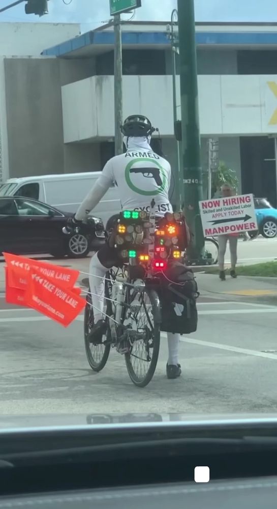 Picture of a cyclist on a bike with at least a dozen lights attached to the back and several flags sticking out from his left saying "take your lane" the cyclist is wearing a tight fitted white cycling short with a picture of a gun printed on the back captioned "armed cyclist"