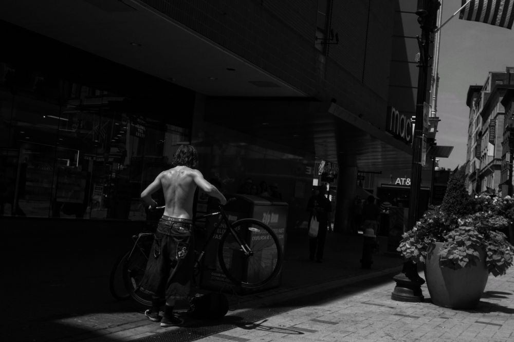 Black and white photo of a shirtless man, seen from behind, lifting a bike off a bike rack. He is in a strip of sunlight and stands out against the shadows. To the right of the frame is a large planter and distant buildings in the sun.