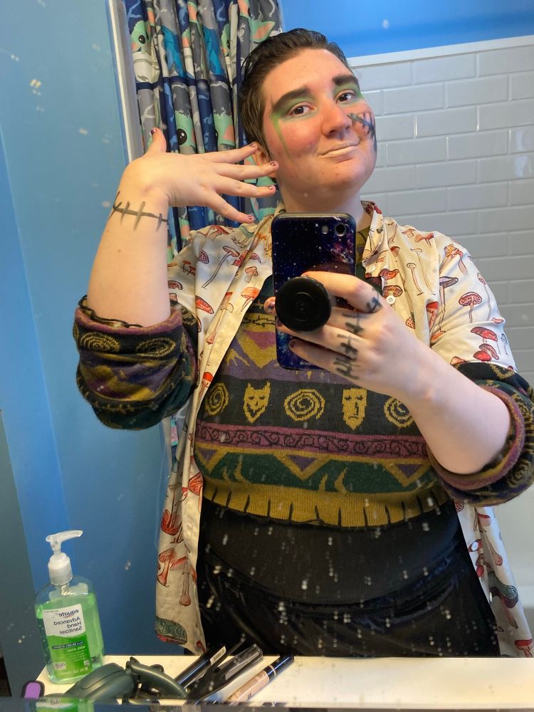 Frankenstein's Monster inspired makeup to go see Guillermo del Toro's Frankenstein 
An androgynous 20 something with green and silver eyeshadow, pale concealer used as lipstick, and a metallic black stitch drawn on their left cheek with lipstick takes a mirror selfie. They are wearing a tan Hawaiian shirt with red mushrooms on it, a Nightmare Before Christmas crop sweater over a black tank top, and black athletic shorts. They gesture with their right hand towards their face and makeup and take the selfie with their phone in their left hand. They have drawn on stitches on their right wrist and the fingers on their left hand. 