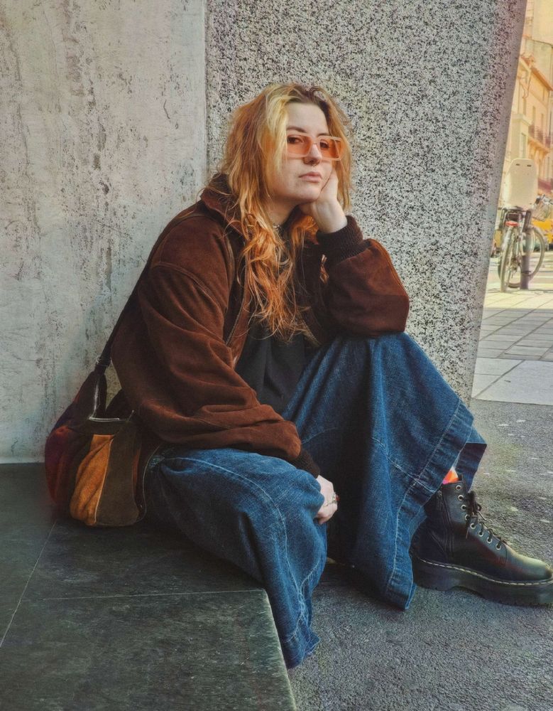 Photo de moi assise dehors: Je porte une jupe-culotte en jean, un bomber en daim marron, des Doc Martens noires à plateformes, et des lunettes de soleil rosées