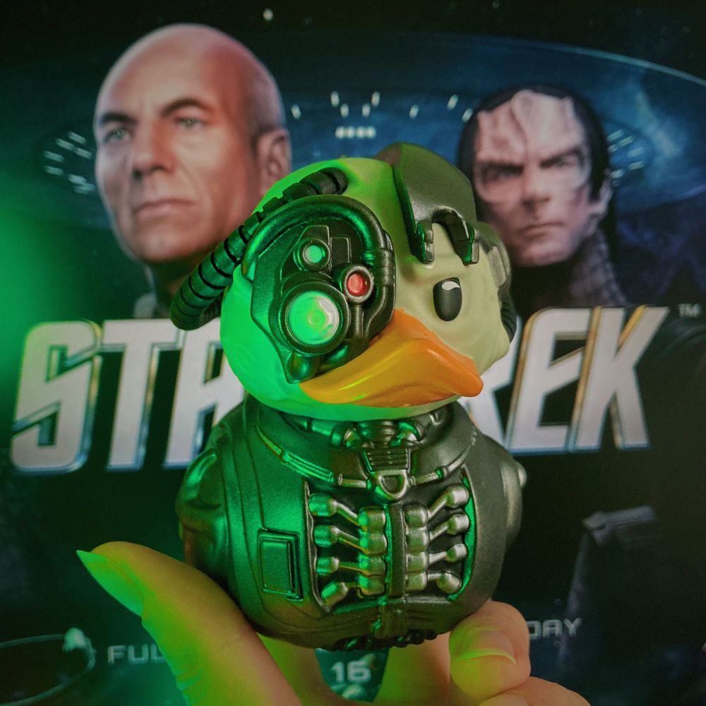 A rubber duck in the style of Jean-Luc Picard from Star Trek, wearing a Locutus of Borg cybernetic costume, is being held up by an index finger and a thumb. It is placed in the centre of the image in front of a computer screen background - a blurry version of the key art from Star Trek: Infinite, the 4X grand strategy game.