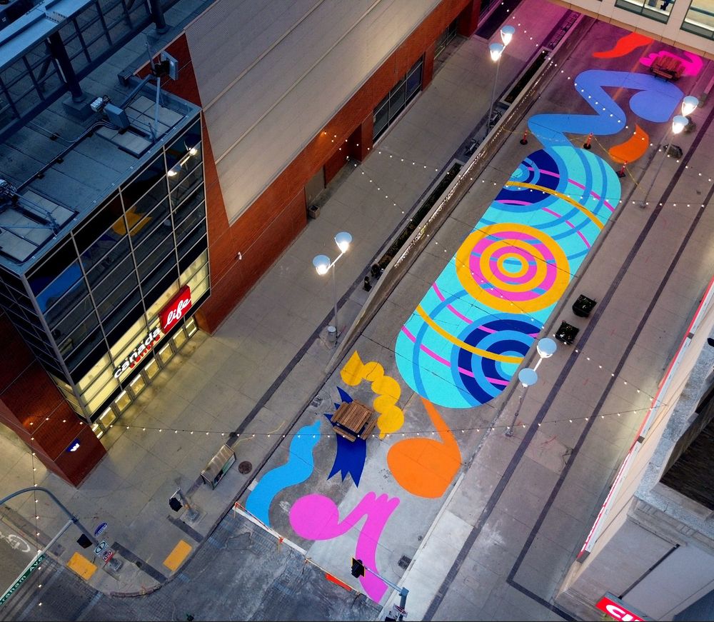 drone photo of graham ave. in winnipeg next to Canada Life Centre: a skateboard-shaped ground mural with a design of overlapping concentric circles in bright colours, surrounded by various whimsical and bouncy shapes painted on the rest of the road.