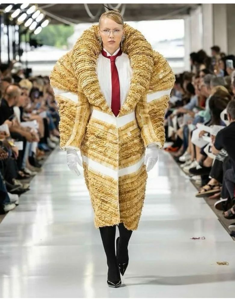 A model wearing a yellow and white puffy outfit like a skirt/ swear combo, with a large collar.  She has a white shirt and a red tie underneath. She has black pantyhose and black heels on. 