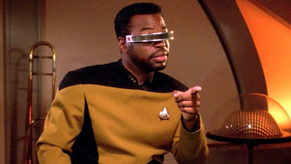 Levar Burton as Georgi LaForge in Star Trek, the Next Generation. He is wearing a gold and black uniform and a VISOR over his eyes, and point at the camera